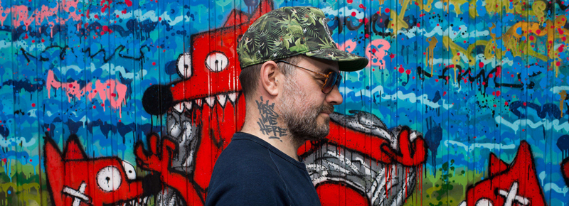 Vue de profil de PIMAX artiste Street-art . En arrière plan une fresque représentant Nourf, son alter ego artistique.