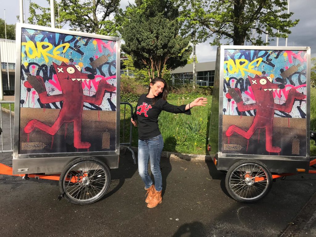Promotrice entouré de 2 vélos publicitaires par une fin d'après-midi ensoleillée. BELAIR FINEART a contacté MediaShift pour un partenariat autour de Pimax artiste de Street-art