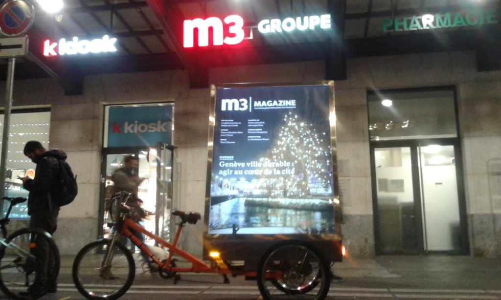 Vélo publicitaire électrique MediaShift avec une affiche m3 magazine devant les locaux de m3 GROUPE